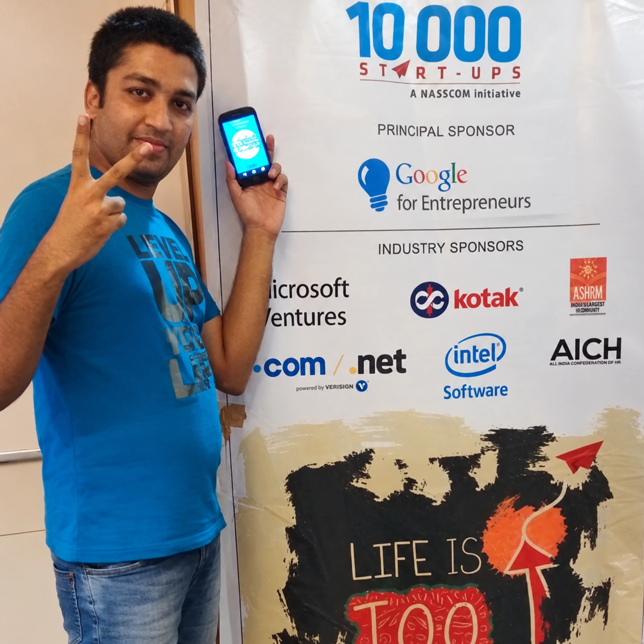Enhanced archive photo of Sumesh beside a 10,000 Startups and Google for Entrepreneurs sponsor board