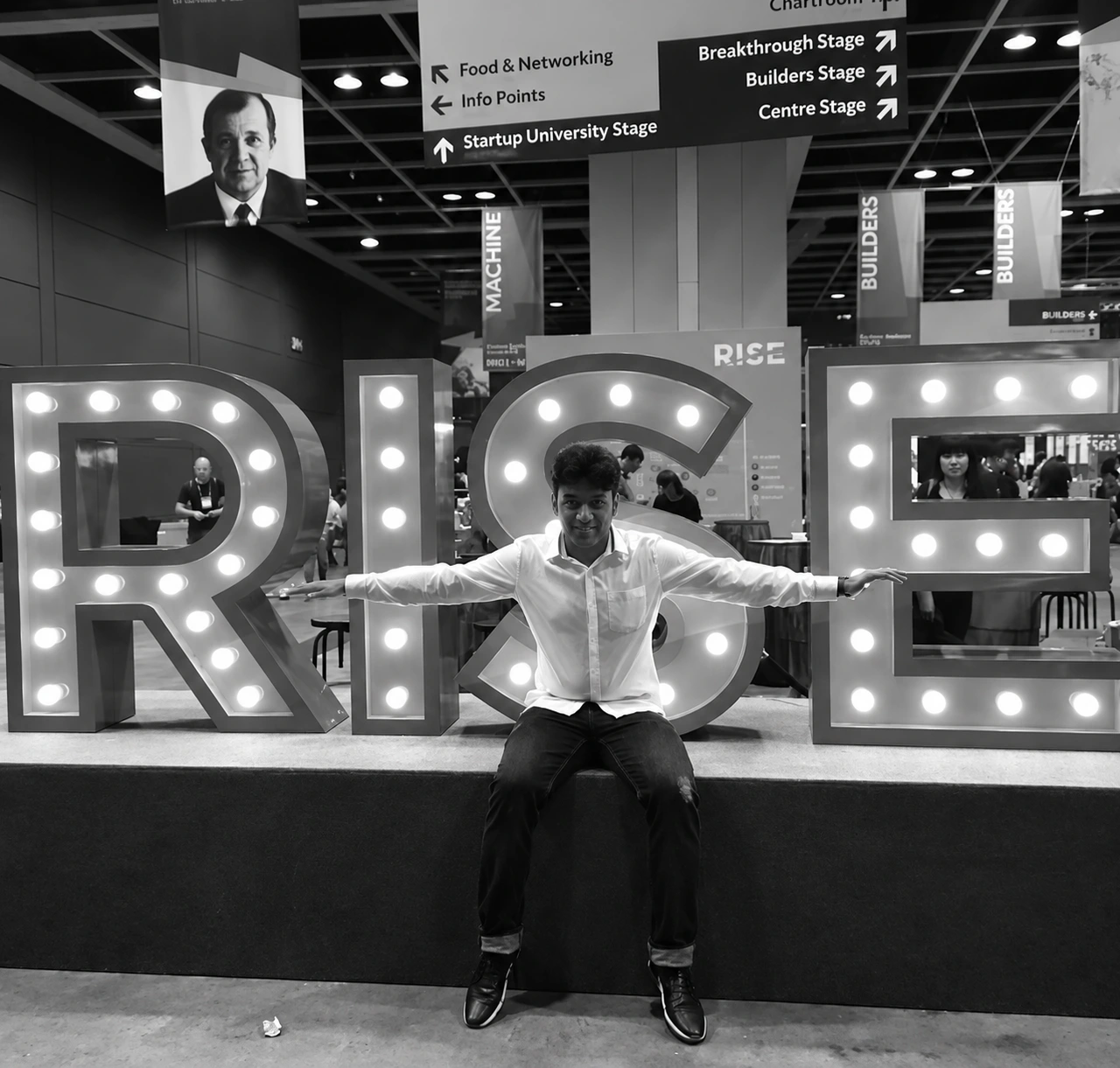 Enhanced black-and-white archive photo of Sumesh seated in front of the RISE conference sign